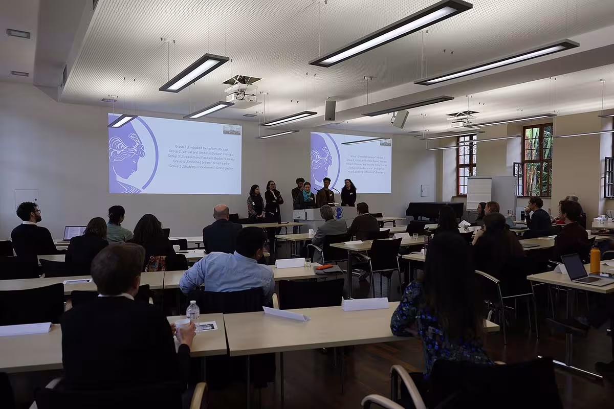 Une salle de conférence. Au premier plan, plusieurs rangées de personnes sont assises. À l'arrière-plan, un groupe se tient devant un pupitre, une présentation est projetée sur le mur.