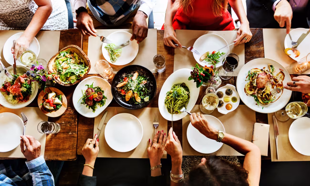 Une table dressée vue d'en haut. Au centre se trouvent des plats issus de différentes cuisines culturelles. À l'extérieur, coupés de l'image et reconnaissables uniquement à leurs bras, les convives sont en train de se servir divers plats dans leurs assiettes.